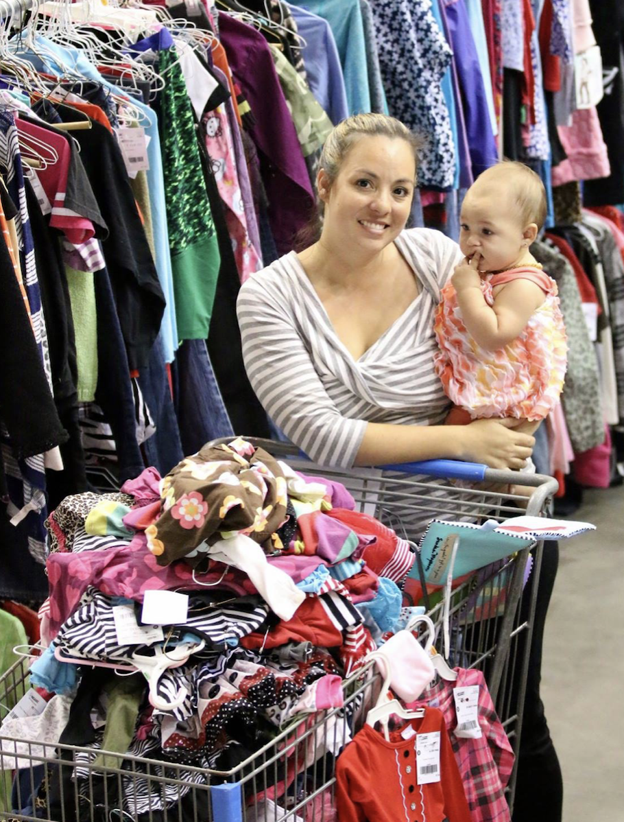A mom holding her baby with a large, overflowing shopping cart at a JBF sale.